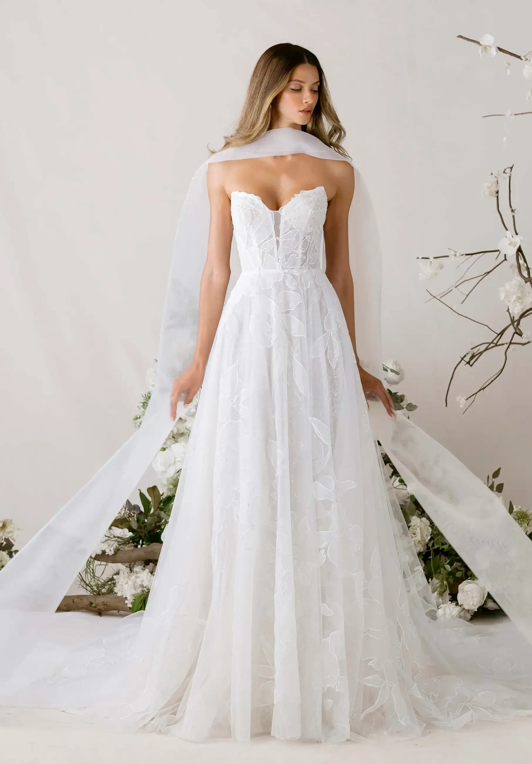 A woman in an elegant, strapless white wedding dress with floral patterns stands among white flowers and branches. The mood is serene and graceful.