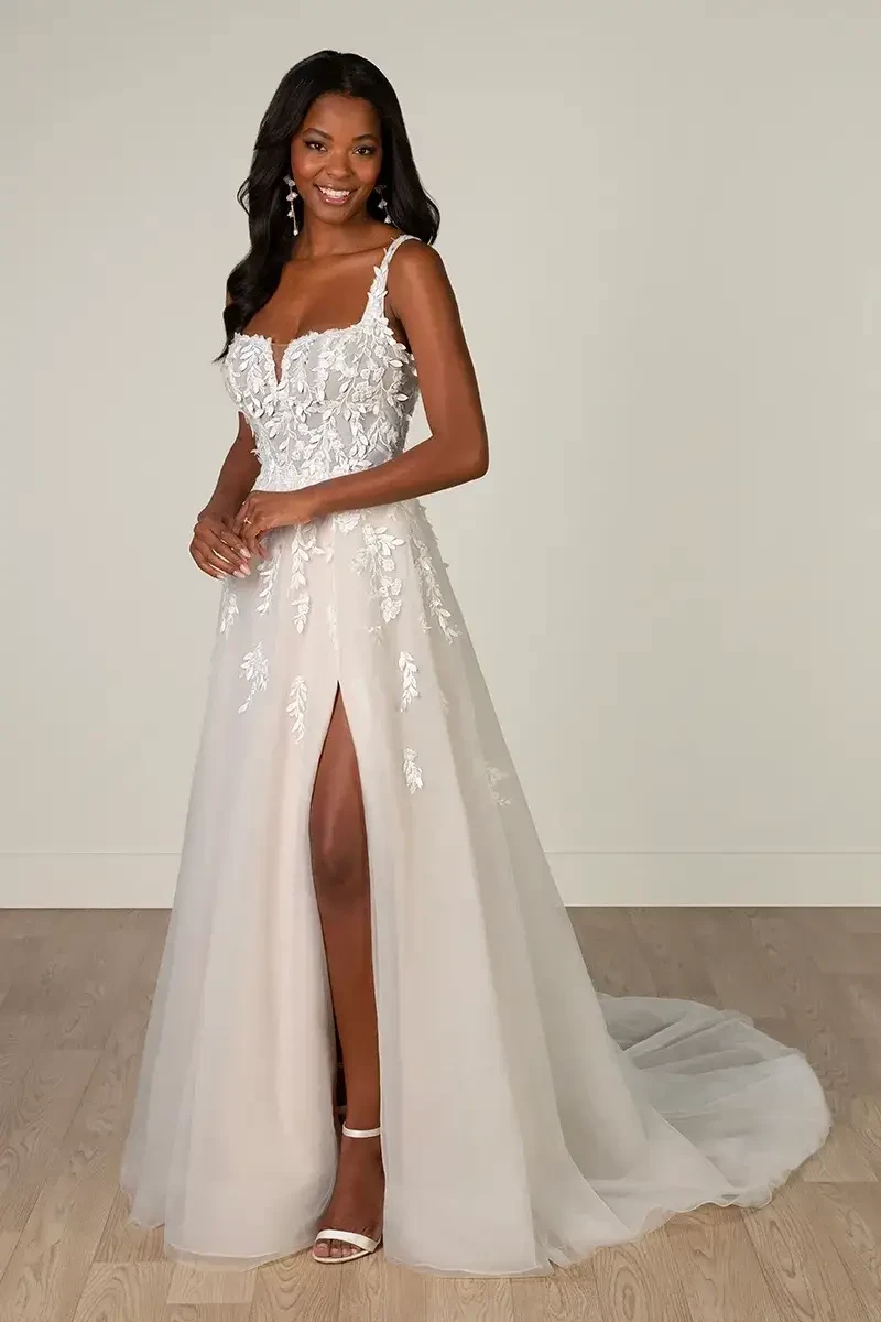 Model is wearing a soft A-line wedding dress featuring a square neckline with a sheer plunge, delicate straps, 3D floral lace appliqués, and a high leg slit.