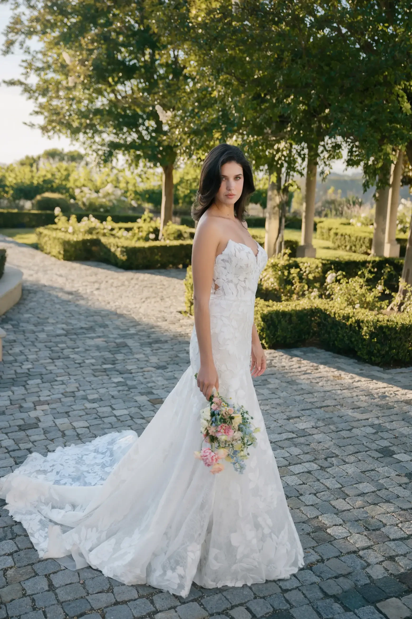 Bride in a white lace strapless gown holds a bouquet, standing on a cobblestone path in a lush garden, exuding elegance and serenity.