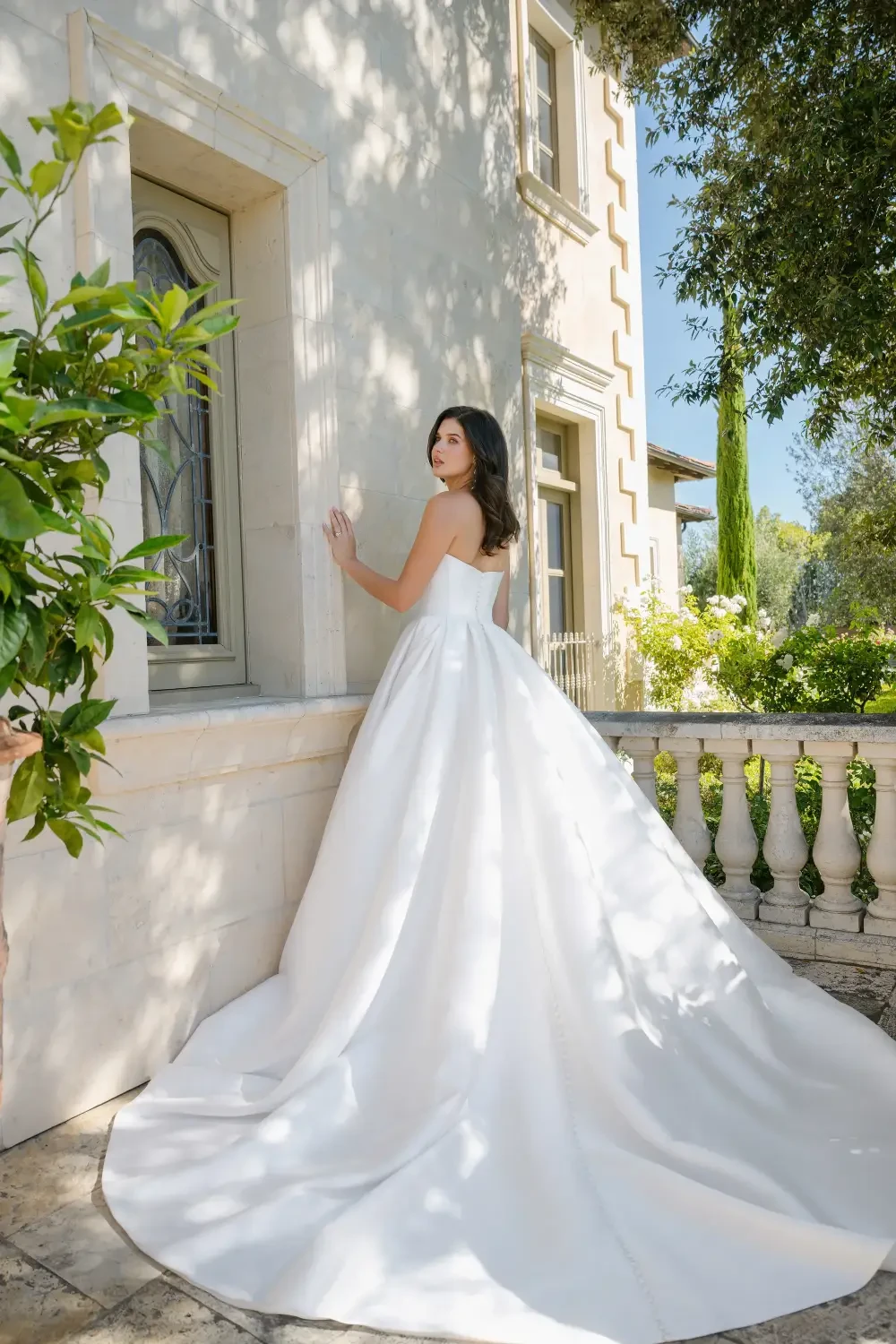 A woman in a stunning white wedding dress stands gracefully beside a building, with lush greenery and sunlight enhancing the romantic setting.