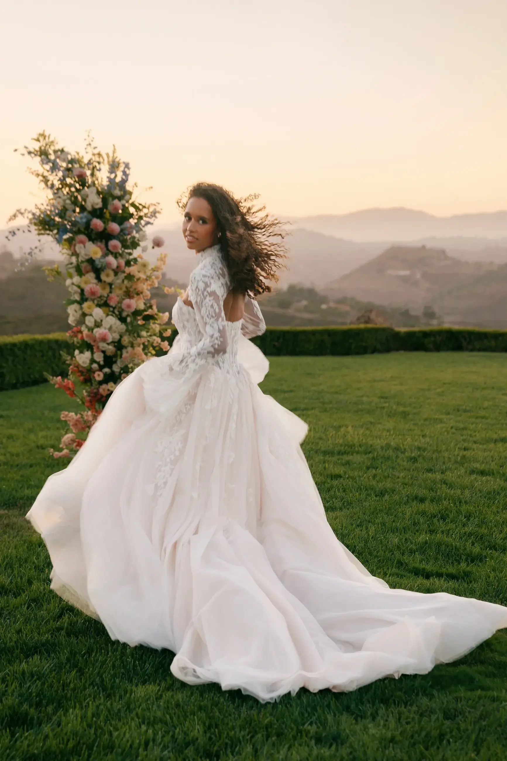 A person wearing a wedding dress looking back while standing on green grass with a floral arrangement in the background and mountains in the distance during sunset.