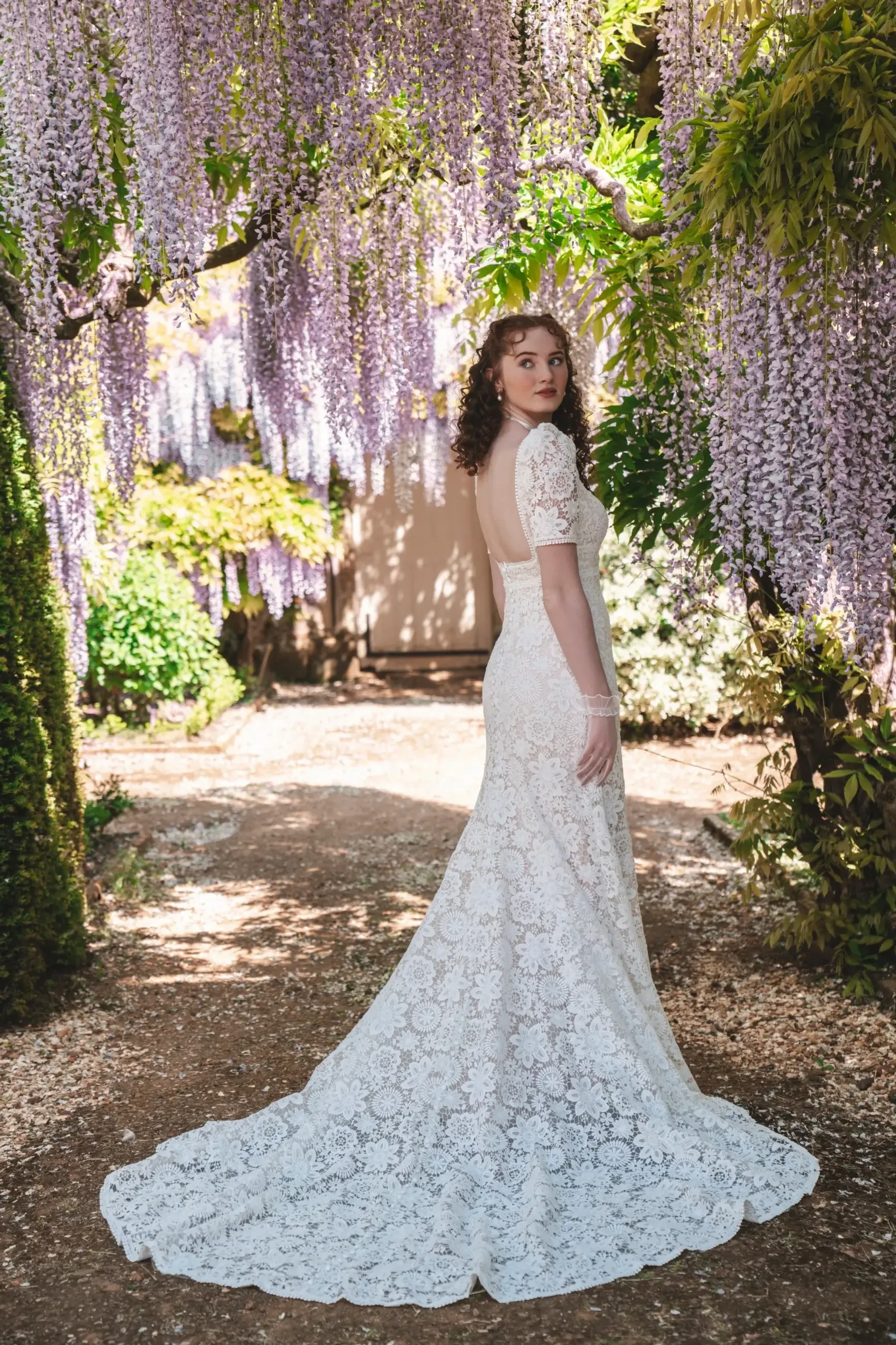 Model is wearing a demure lace sheath gown with short puff sleeves and a scalloped hemline, inspired by vintage regency style.