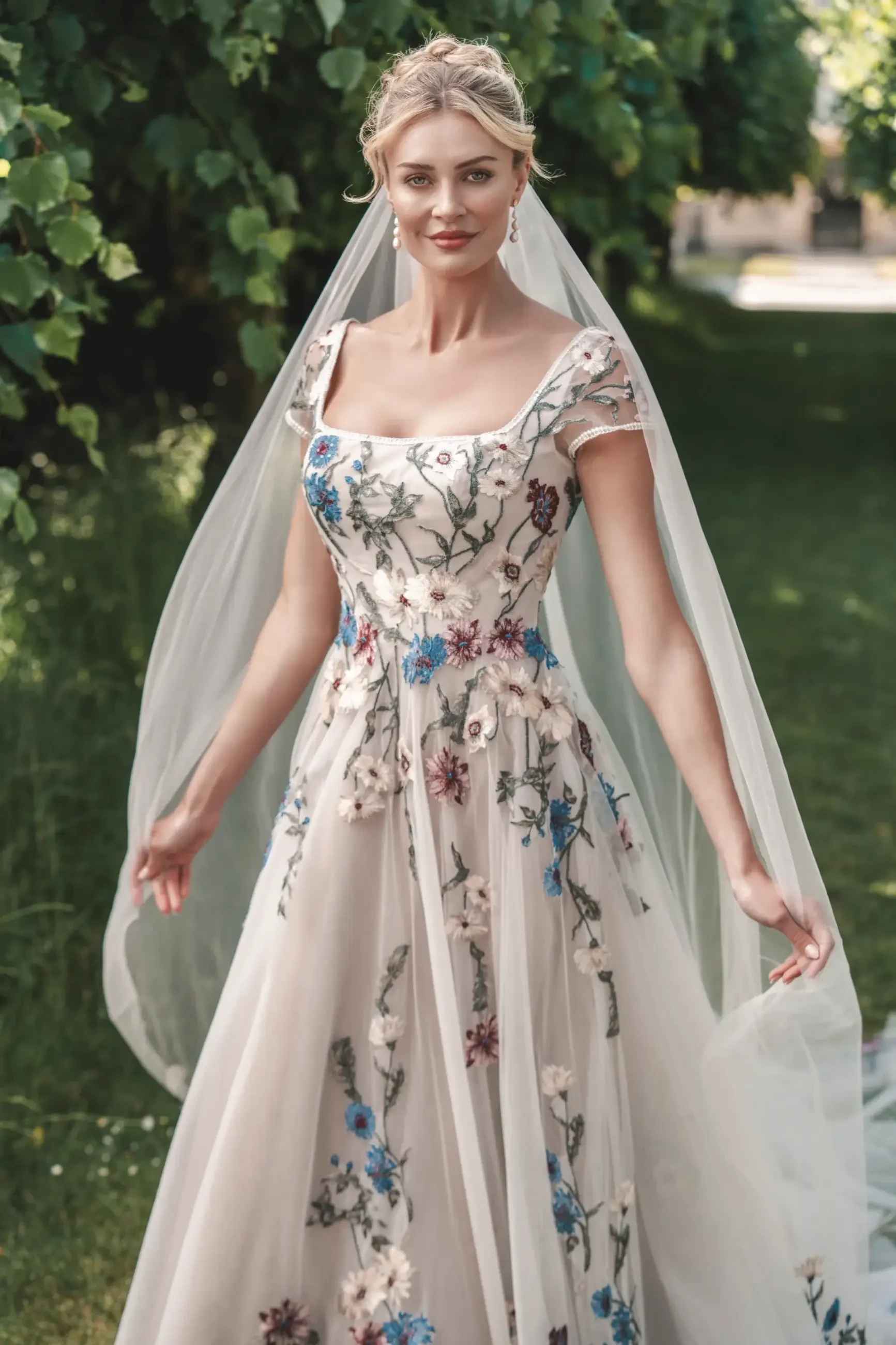 Model is wearing an embroidered tulle A-line gown with ivory, mauve, and blue floral lace and detachable bishop sleeves accented at the wrists.