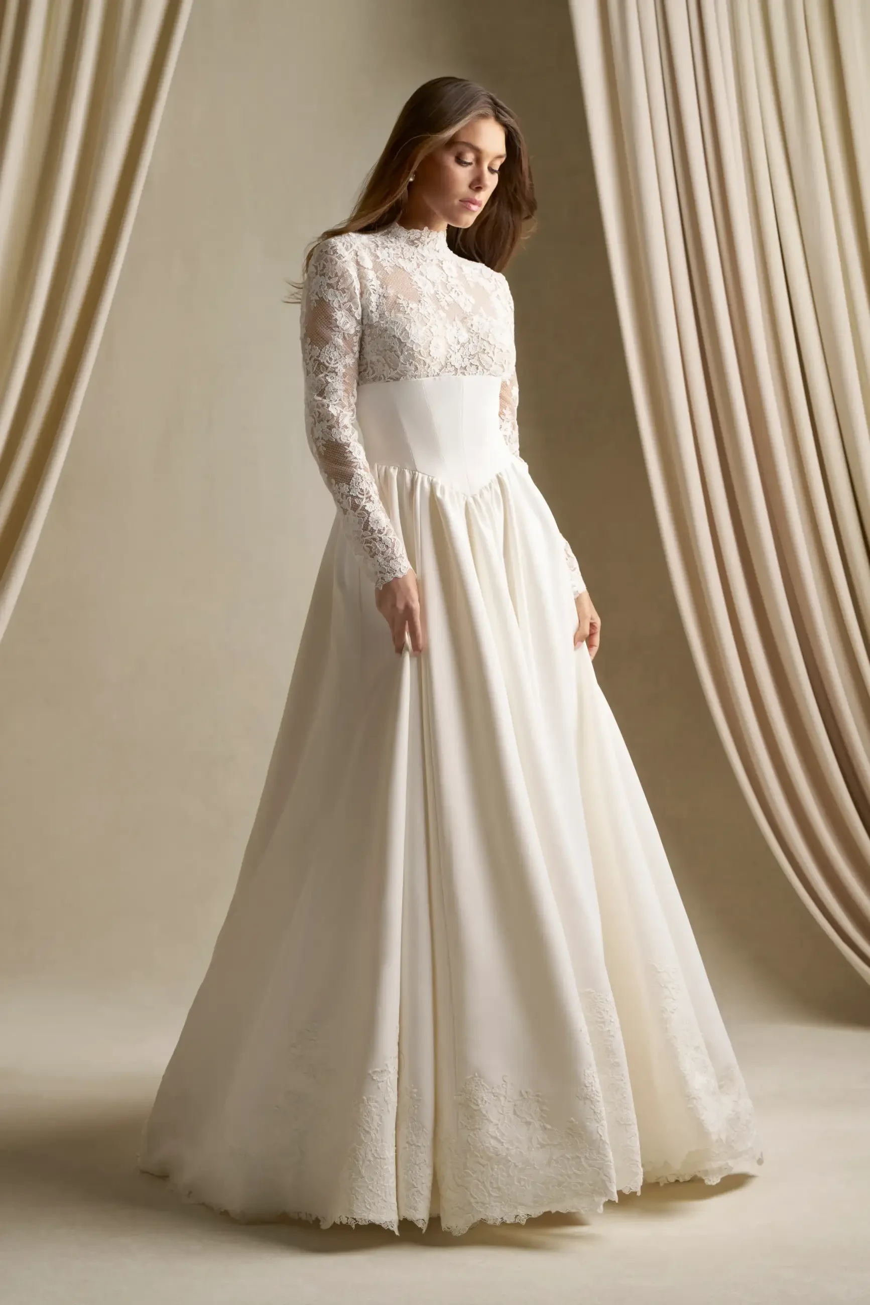 A model wearing a long-sleeved, white wedding dress with lace detailing, standing elegantly against a soft, neutral background with draped fabric.