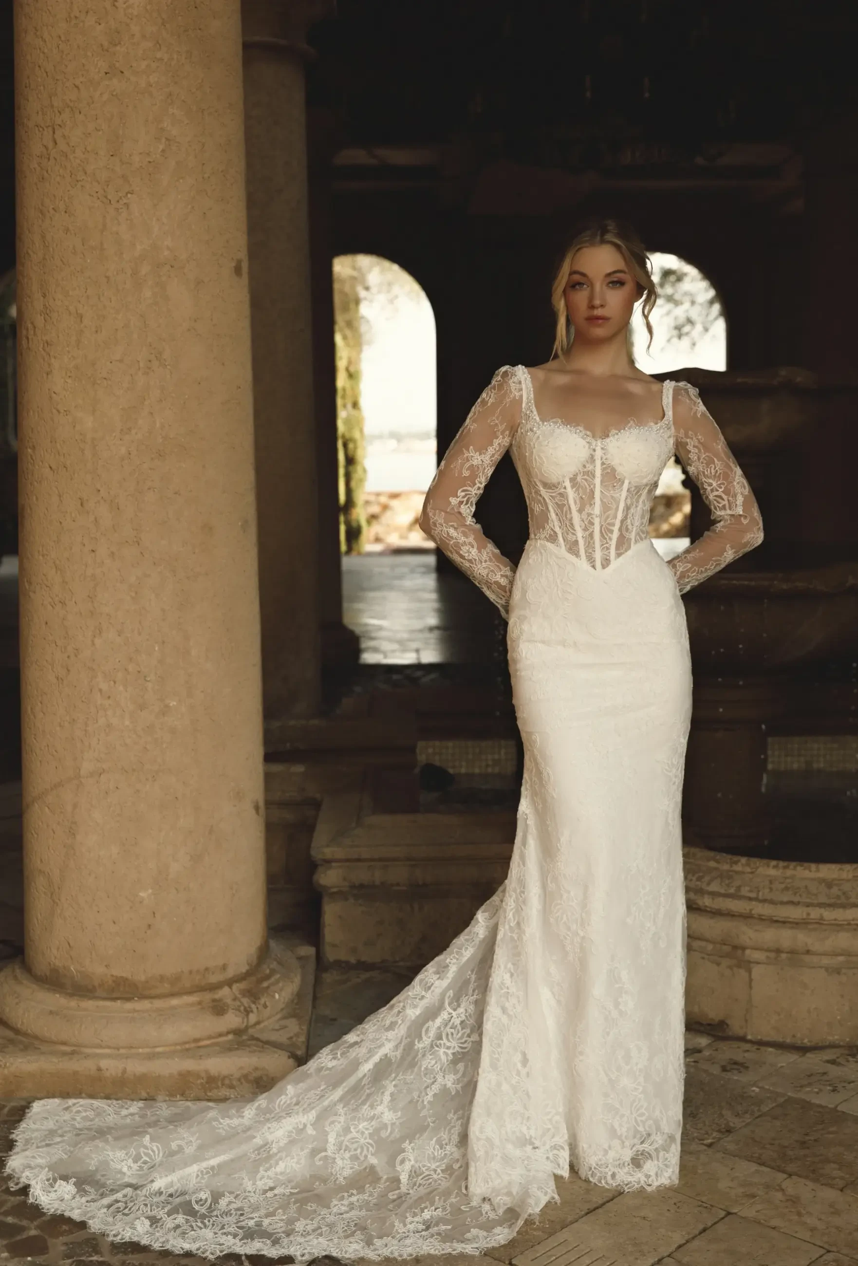 A woman stands in an elegant, form-fitting lace wedding dress with sheer long sleeves and a long train. She's in a dimly lit, archway venue.
