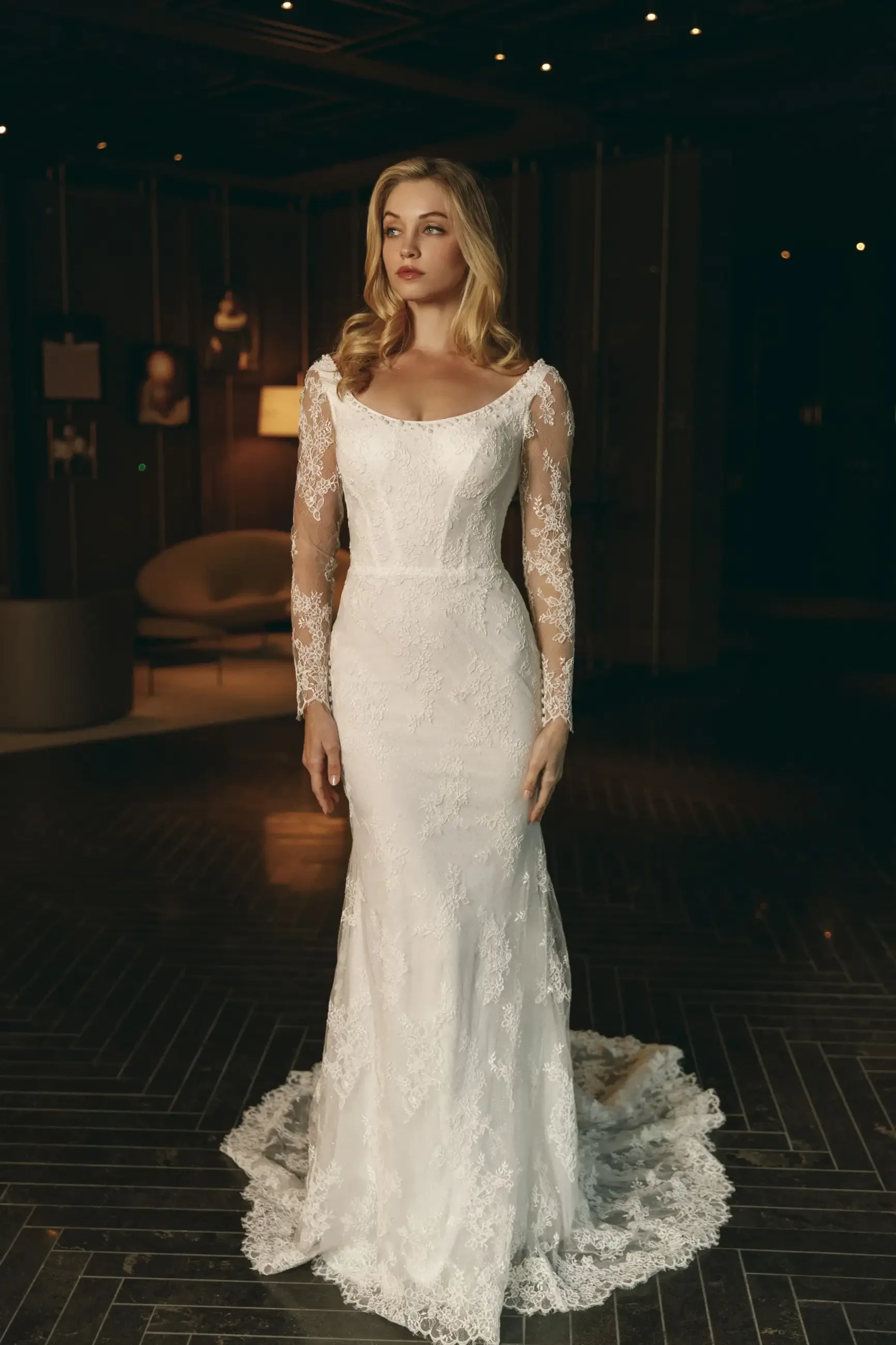 A woman in a long-sleeved lace wedding dress stands elegantly in a dimly lit room.