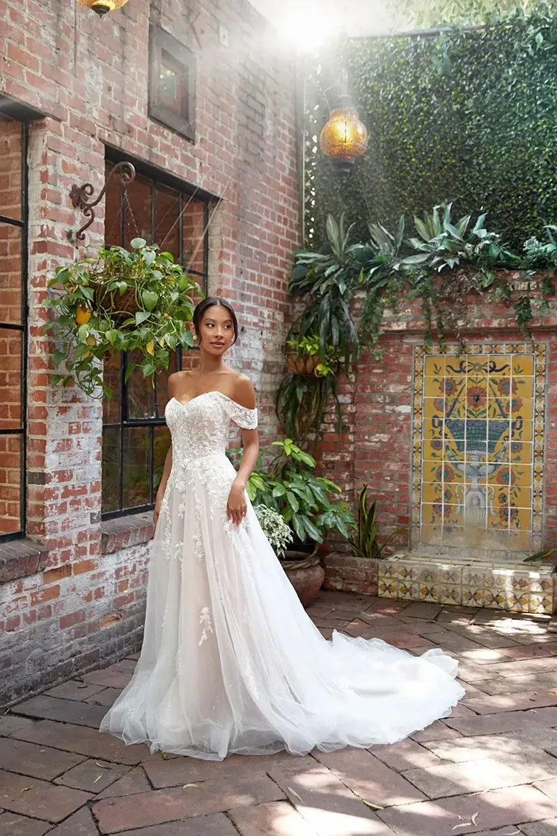 Model is wearing an off-the-shoulder A-line gown with detachable sleeves, a sweetheart neckline, and floral lace appliqués that cascade over glitter tulle.