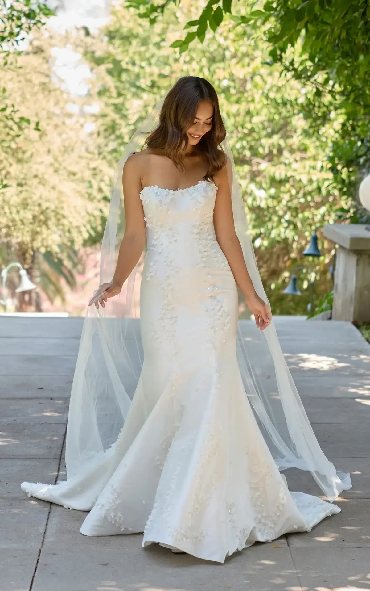 Bride in a strapless, fitted white gown with a long train and veil stands outdoors. She smiles gently, surrounded by lush greenery and soft sunlight.