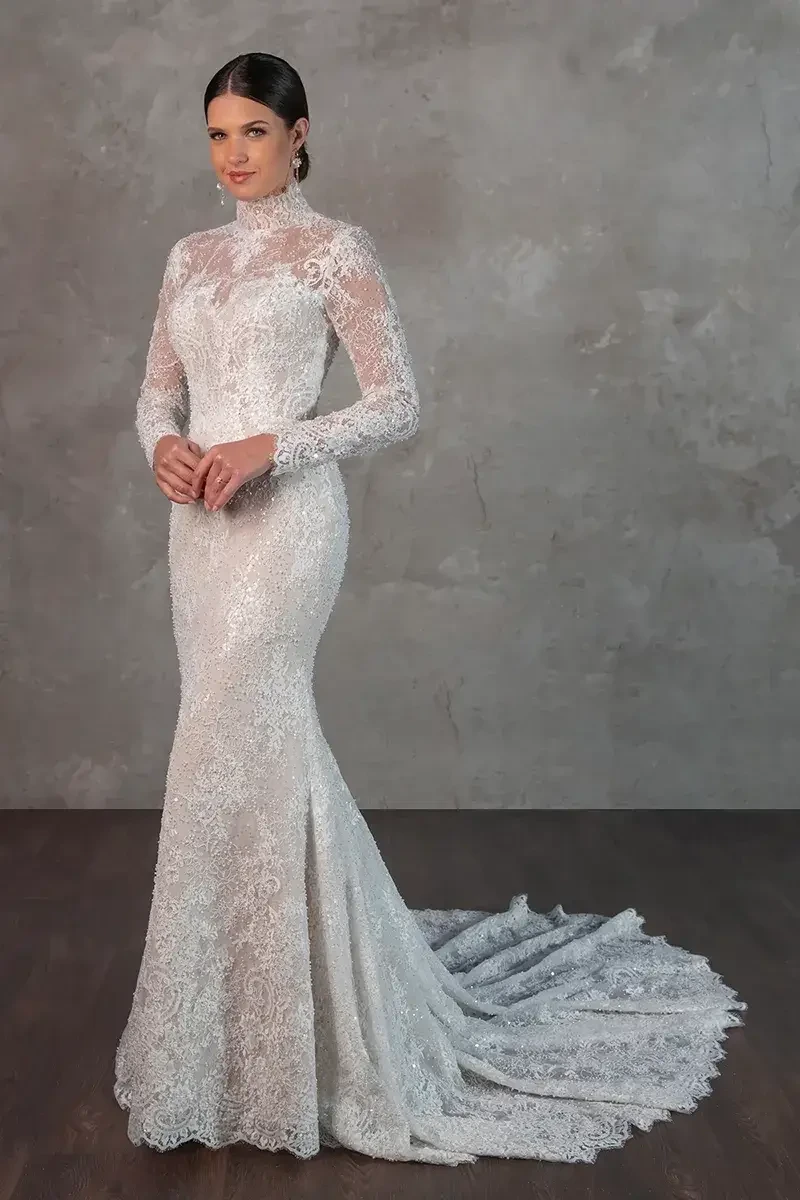Model is wearing a column sheath gown with a sheer all-lace bodice, high mock neckline, beaded Chantilly lace sleeves, fabric-covered buttons, and a sweeping train.