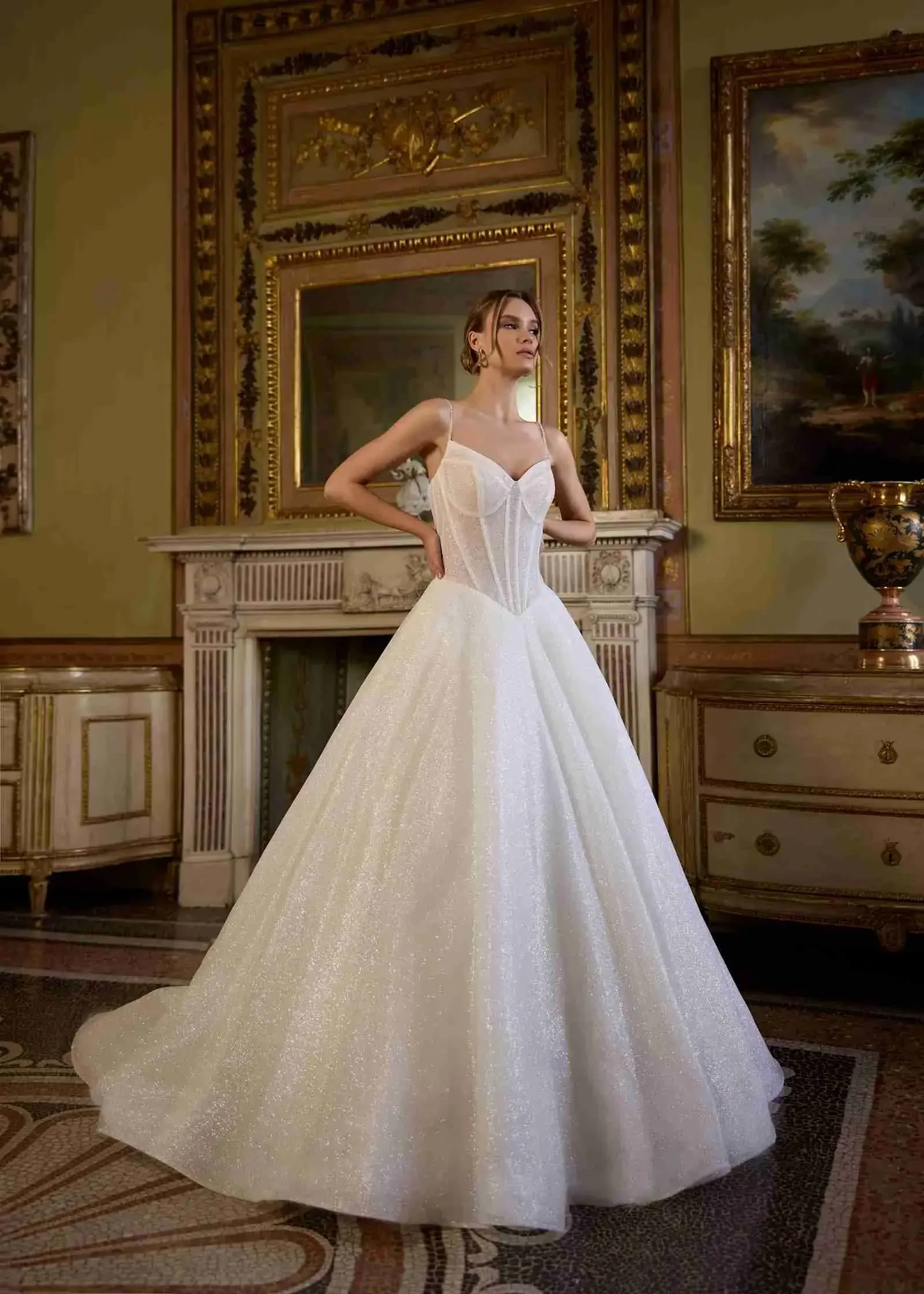 A woman stands elegantly in a shimmering white ball gown with a corset top. The ornate room features a detailed fireplace, vintage decor, and framed art, conveying a sense of grandeur.