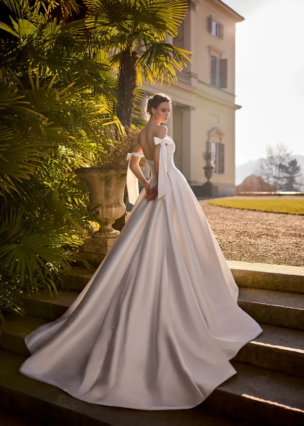 A model in a white wedding gown standing on stone steps surrounded by greenery and a historic building in the background.