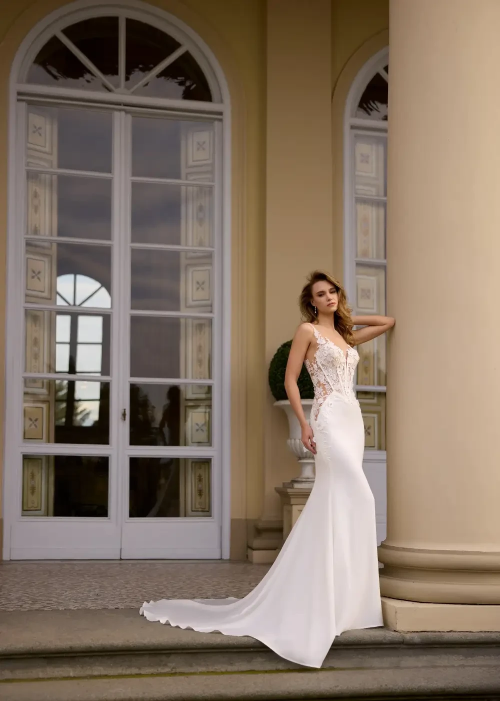 A woman in a stunning white gown poses gracefully by a large window and columns.