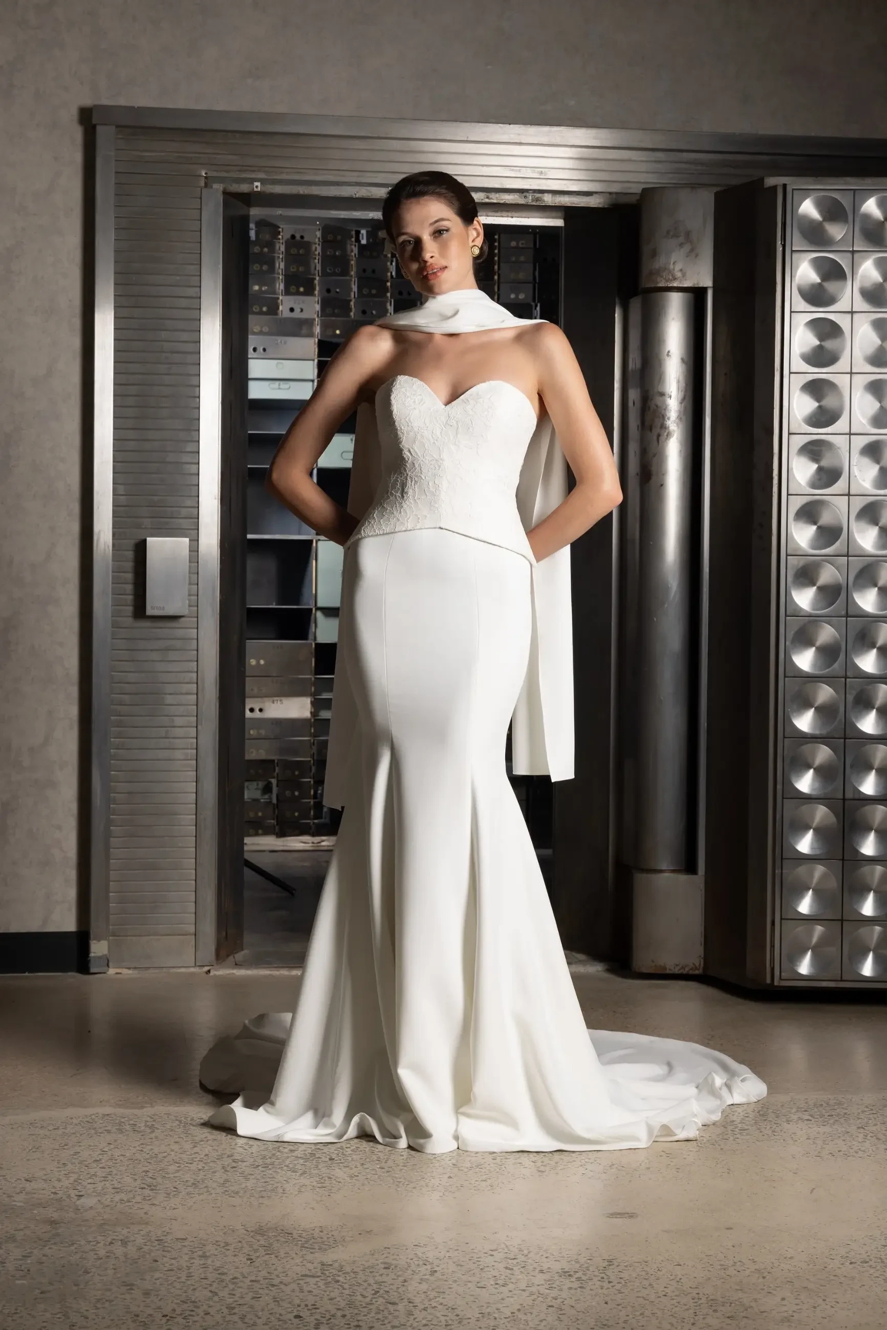 A woman poses confidently in a sleek, strapless white gown with a scarf, standing in front of an open vault door with a modern, industrial backdrop.