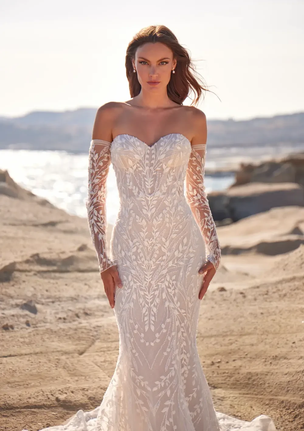 A woman in a strapless, intricate white wedding dress with long sheer sleeves, standing on a rocky shore with the ocean in the background.