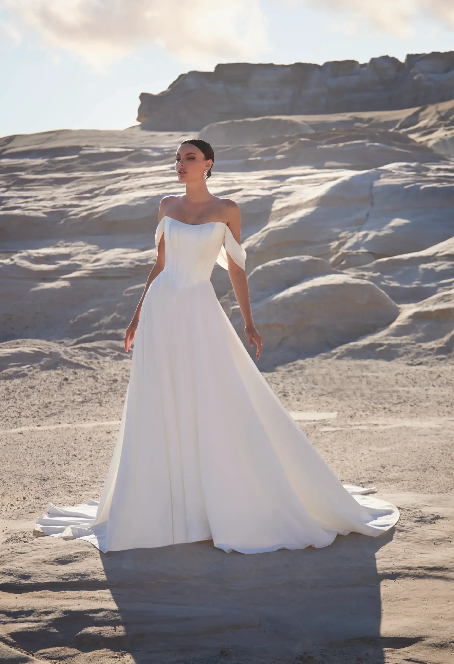 A woman in an elegant white wedding dress stands on a rocky, sunlit landscape. The dress has an off-shoulder design, flowing gracefully around her.