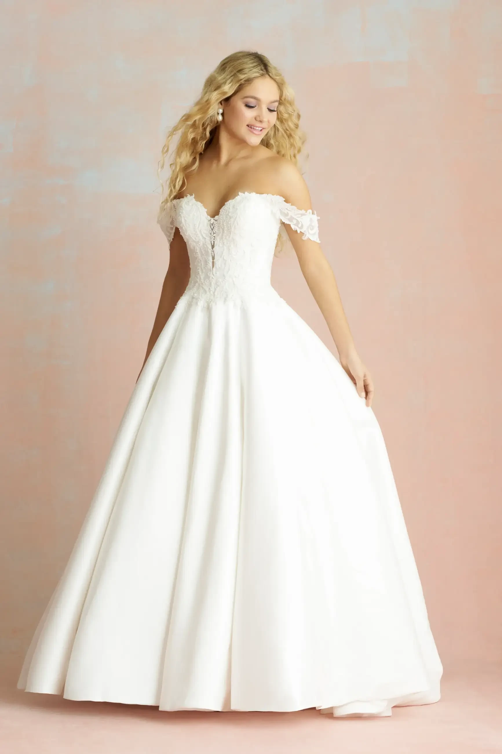 Model is wearing a Mikado ballgown with a plunging sweetheart neckline, off-the-shoulder sleeves, a Basque waistline, pearl lace appliqués, and a sheer back.