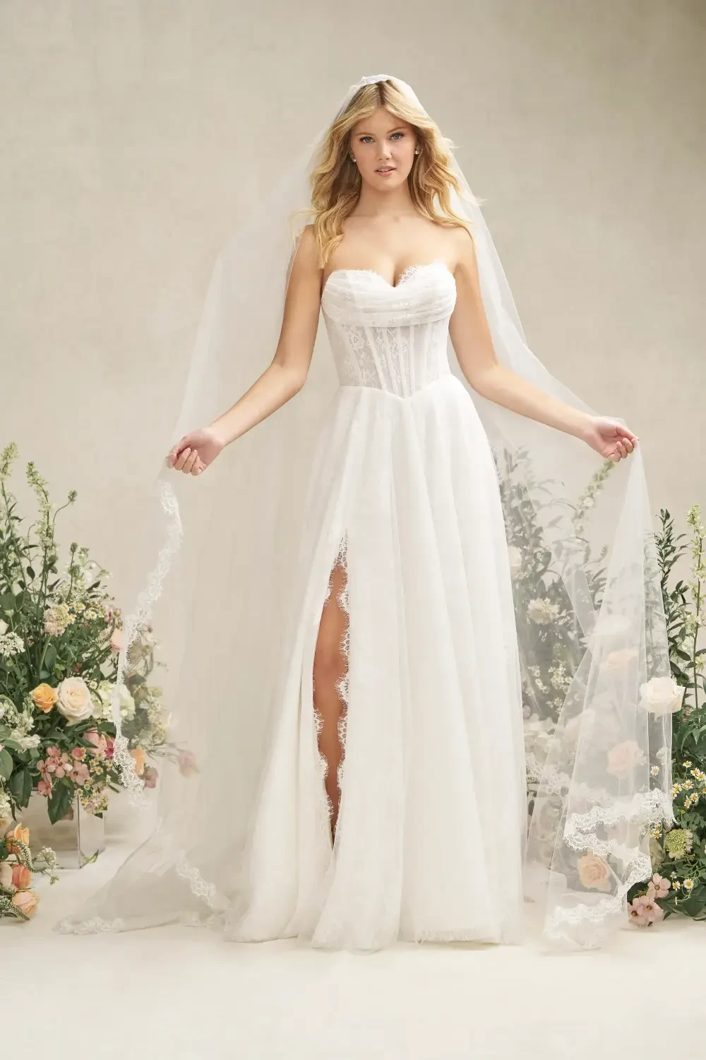 A woman in a white wedding dress with a strapless bodice and a thigh-high slit, holding her veil, surrounded by flowers.