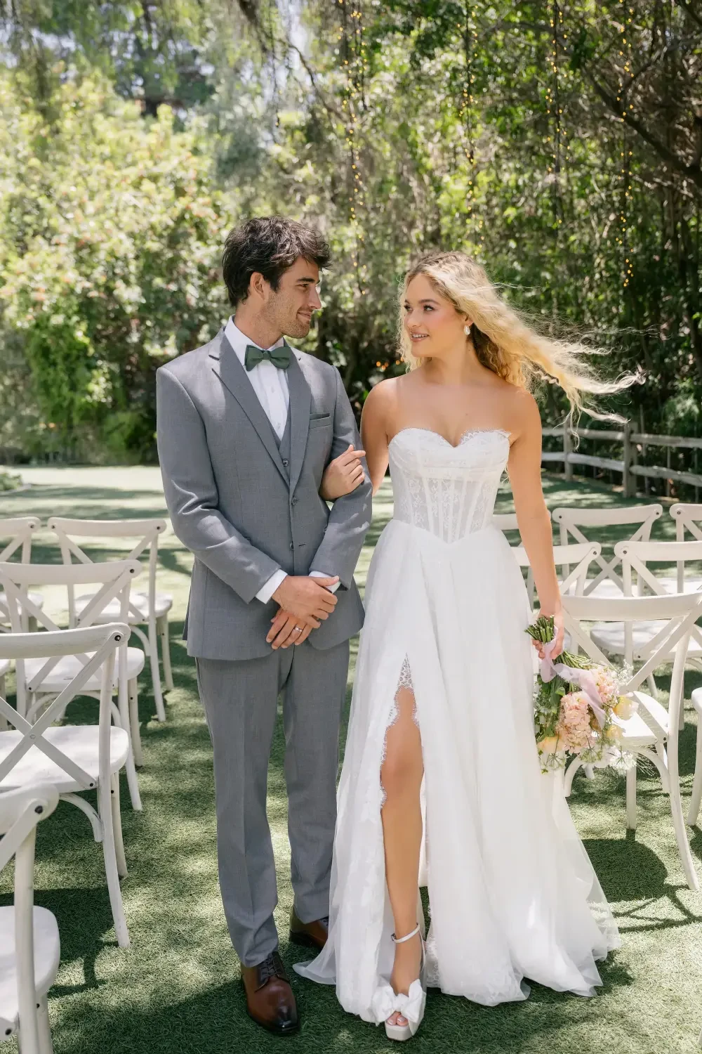 A couple walking together in a scenic outdoor setting, with the woman wearing a white wedding dress and holding a bouquet, and the man in a gray suit with a bow tie.