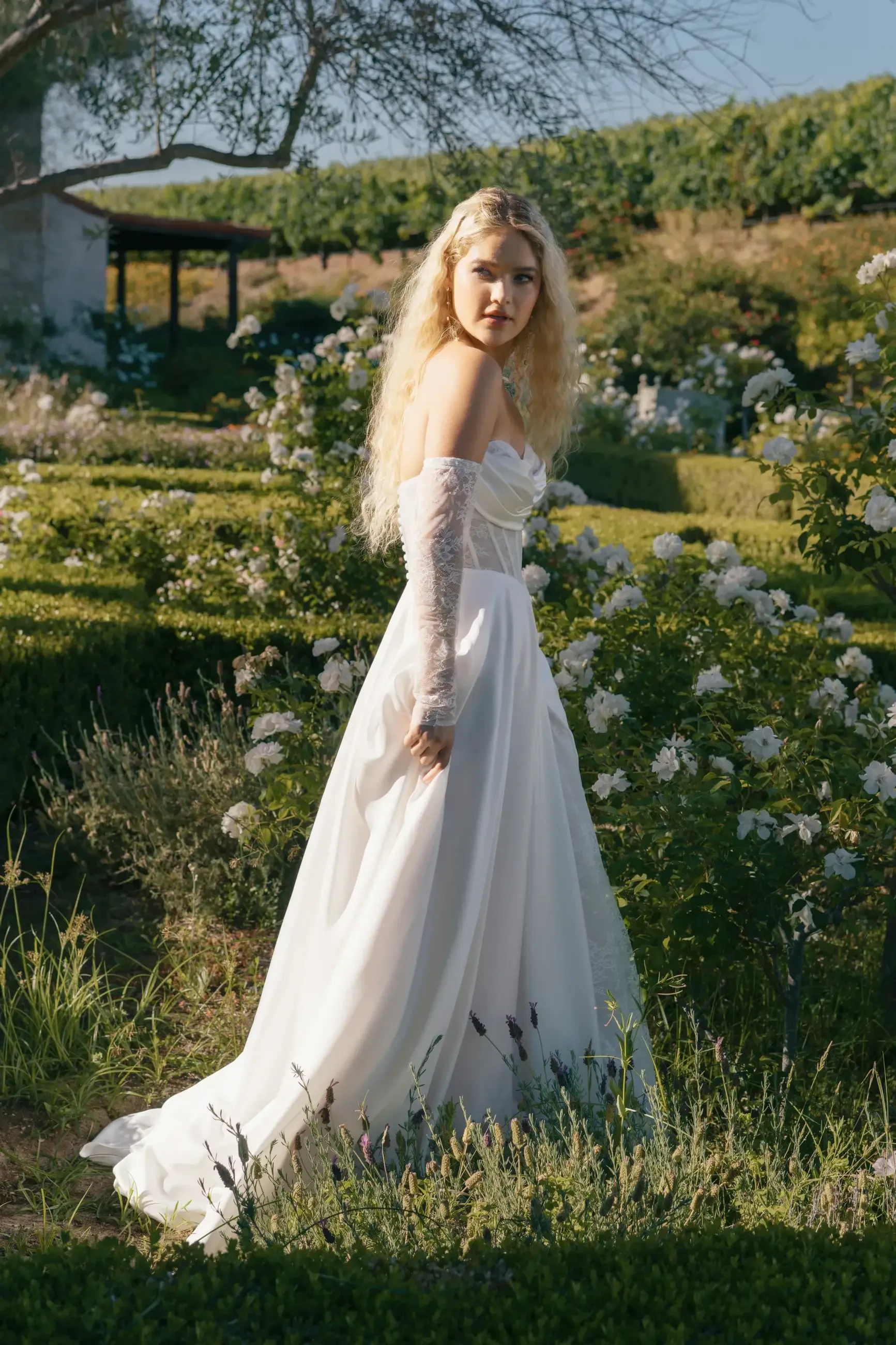 A young woman in a white dress stands in a garden surrounded by flowers, with a serene expression and flowing hair.