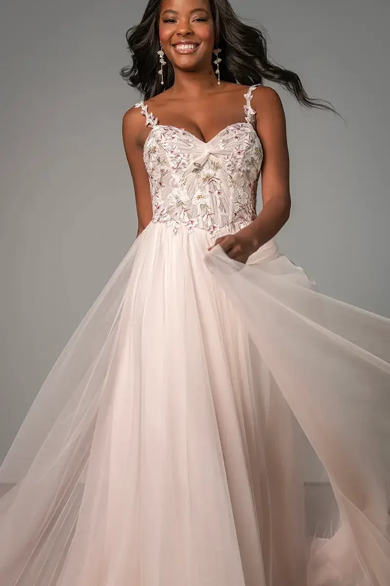 Model is wearing a whimsical A-line gown with embroidered lace, petal-detailed spaghetti straps, a ruched knot bodice, pink tulle layers, and a soft flowing skirt.